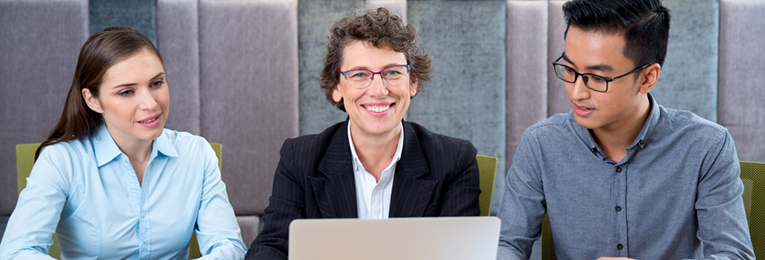 Illustrative image of three people in an office space, with one smiling at the camera while the other two focus on a laptop screen.